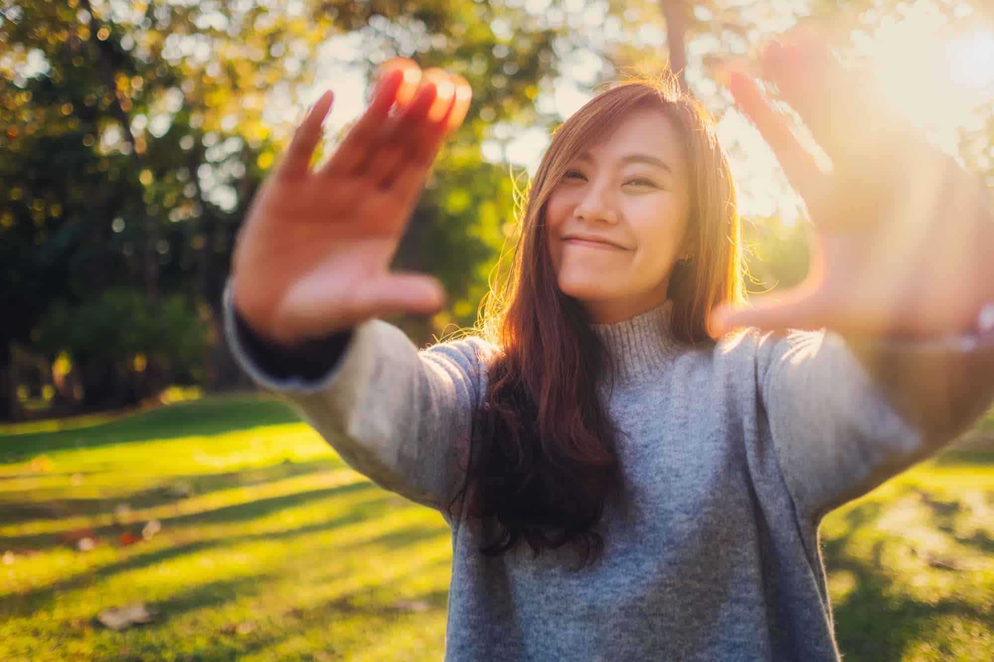 Portrait image of a beautiful asian woman raising hands and playing with camera in park before sunset